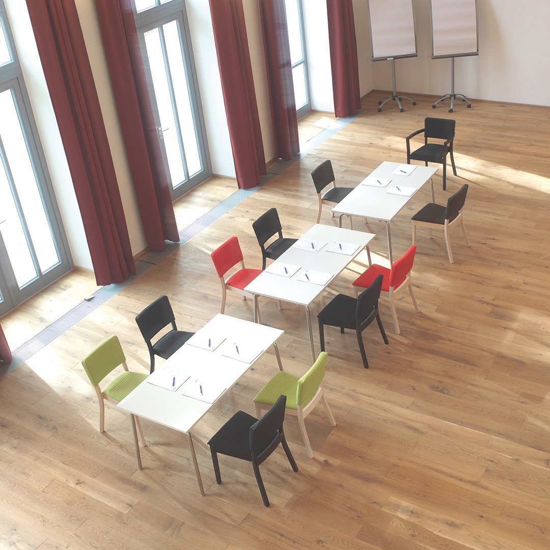 Colorful chairs in red, green, and black paired with white tables in a bright, spacious room with wooden flooring.