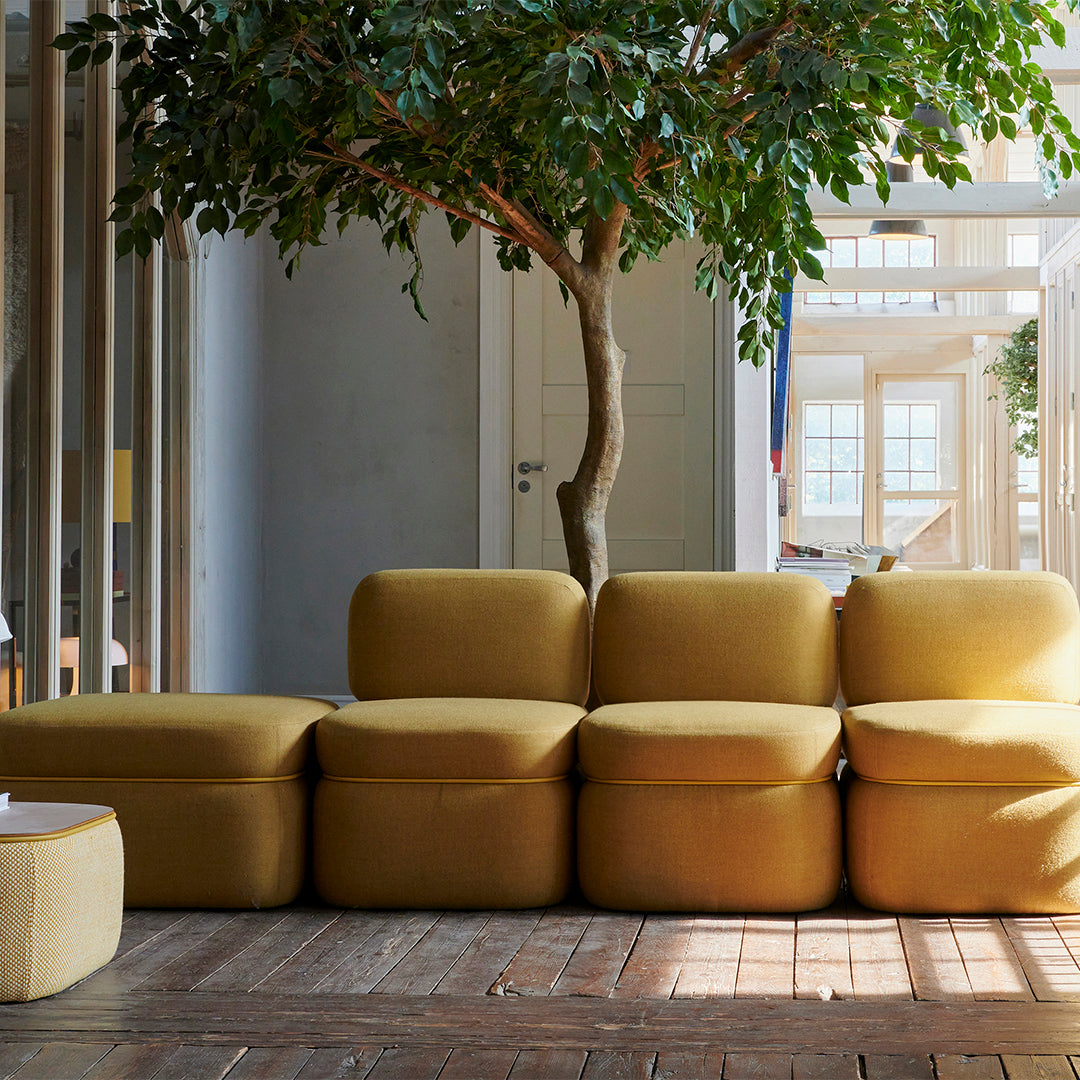 Modern yellow modular sofa with soft, textured fabric against a natural wood floor and greenery backdrop.