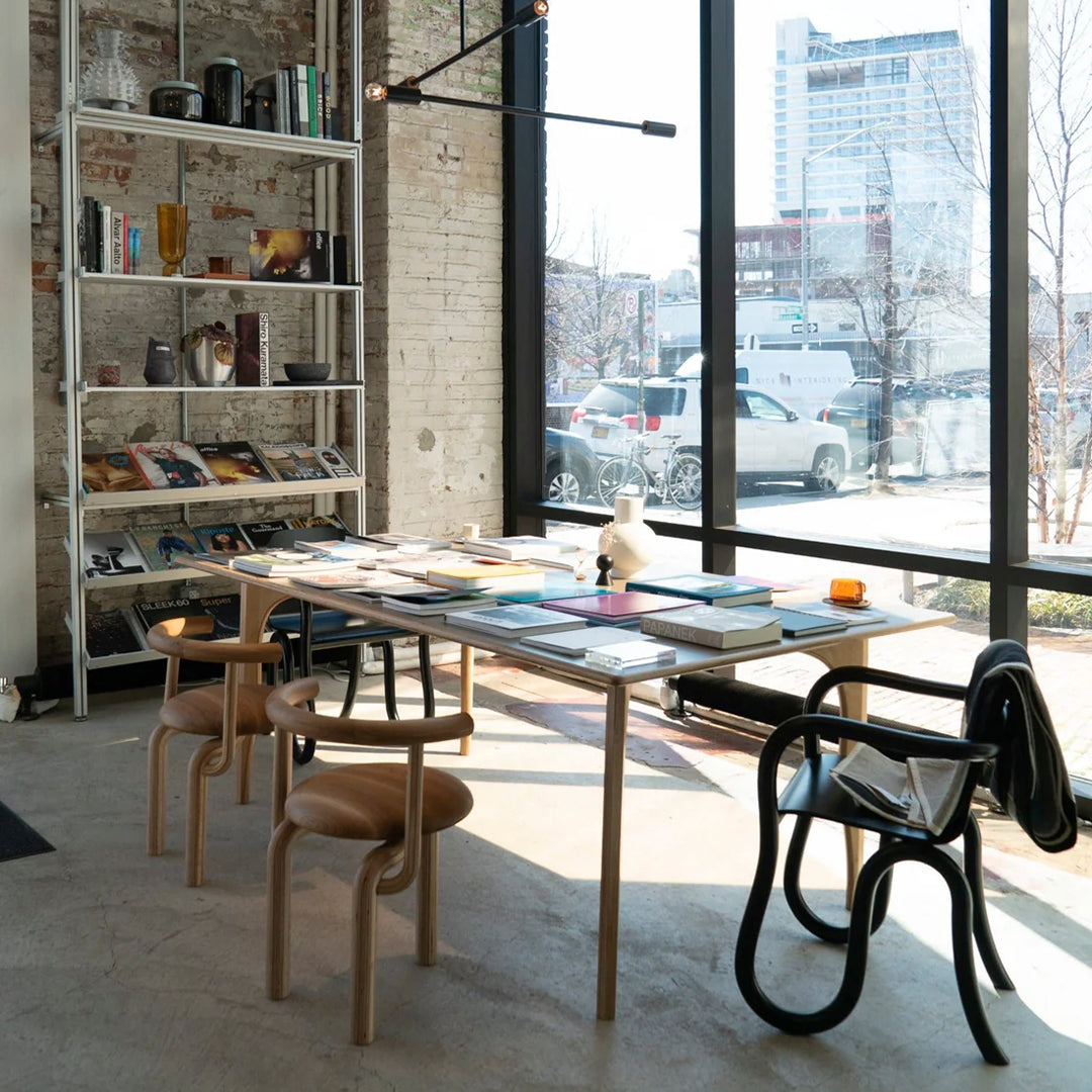 Modern workspace with wooden table, black and natural wooden chairs, vibrant book covers, and neutral brick wall.