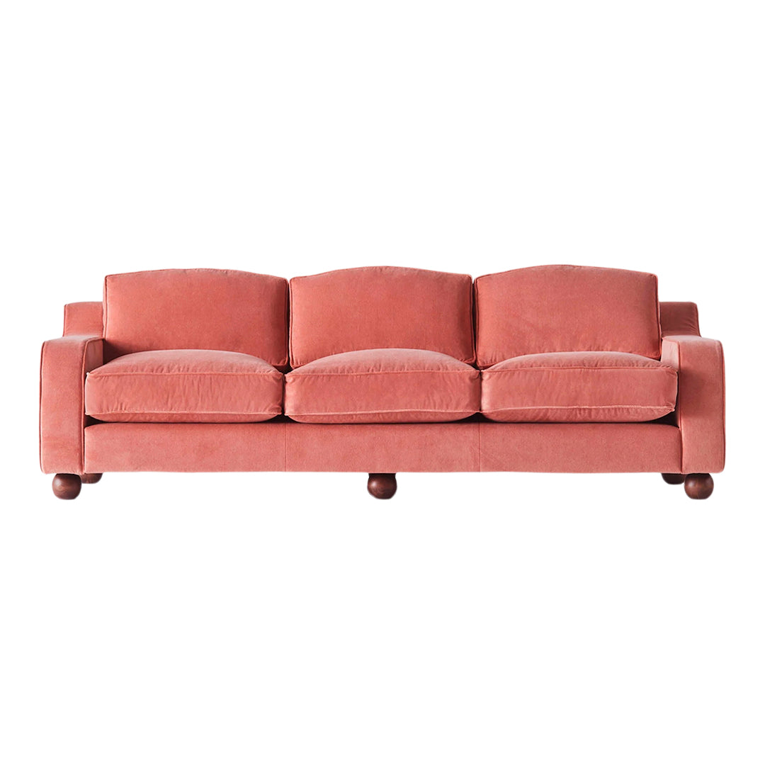 Soft coral velvet sofa with rounded cushions and dark wooden feet. Modern design ideal for cozy living spaces.