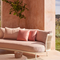 "Modern beige outdoor sofa with soft pink and coral pillows, set against a warm, textured peach wall."