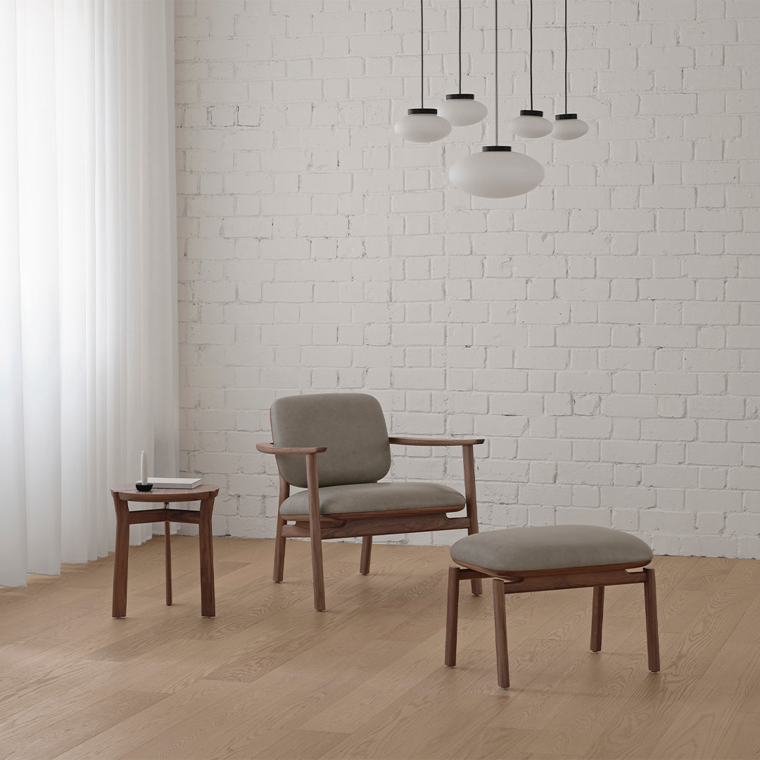 Contemporary gray upholstered chair and ottoman with wooden accents, against a white brick wall and light wood flooring.