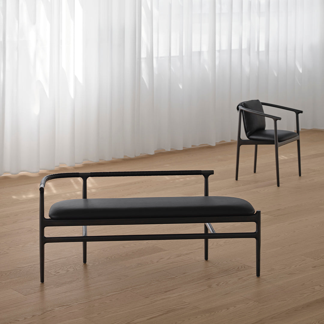 Black metal frame bench with smooth black leather upholstery, set against light wood flooring and sheer white curtains.