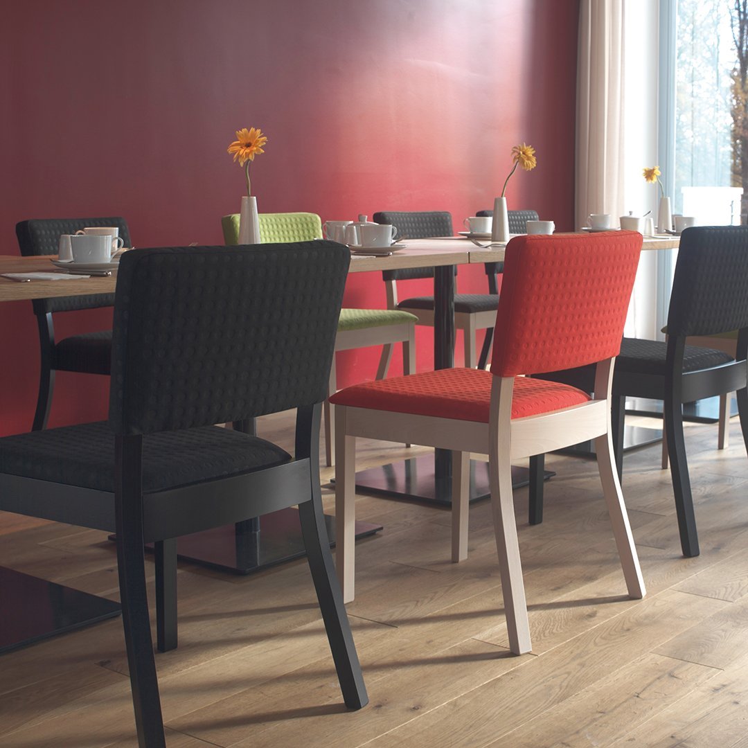 Colorful dining chairs in black, red, green, and cream against a bold red wall, featuring textured patterns and minimalist design.