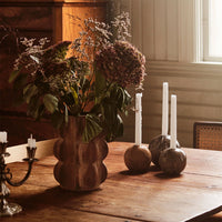 "Mottled brown vase with green foliage, pink hydrangeas, and white candles atop a rustic wooden table."