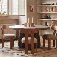 Round concrete dining table with natural wood accents and textured cream chairs, set in a warm, neutral-toned room.