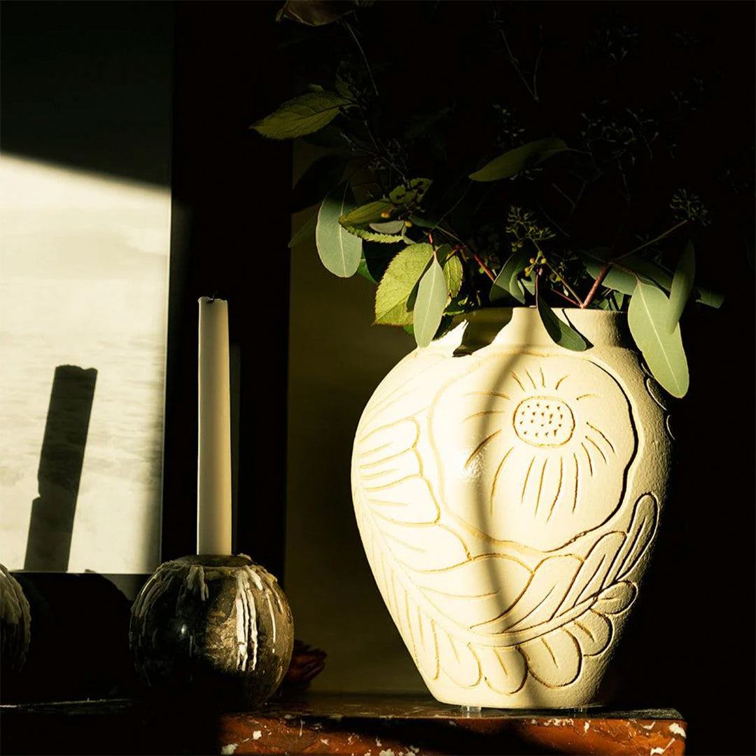 "Decorative cream vase with floral patterns, accented by greenery and sunlight shadows, against a dark background."