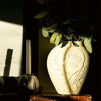 Cream floral-patterned vase with delicate etchings, complemented by lush green foliage and soft shadows.