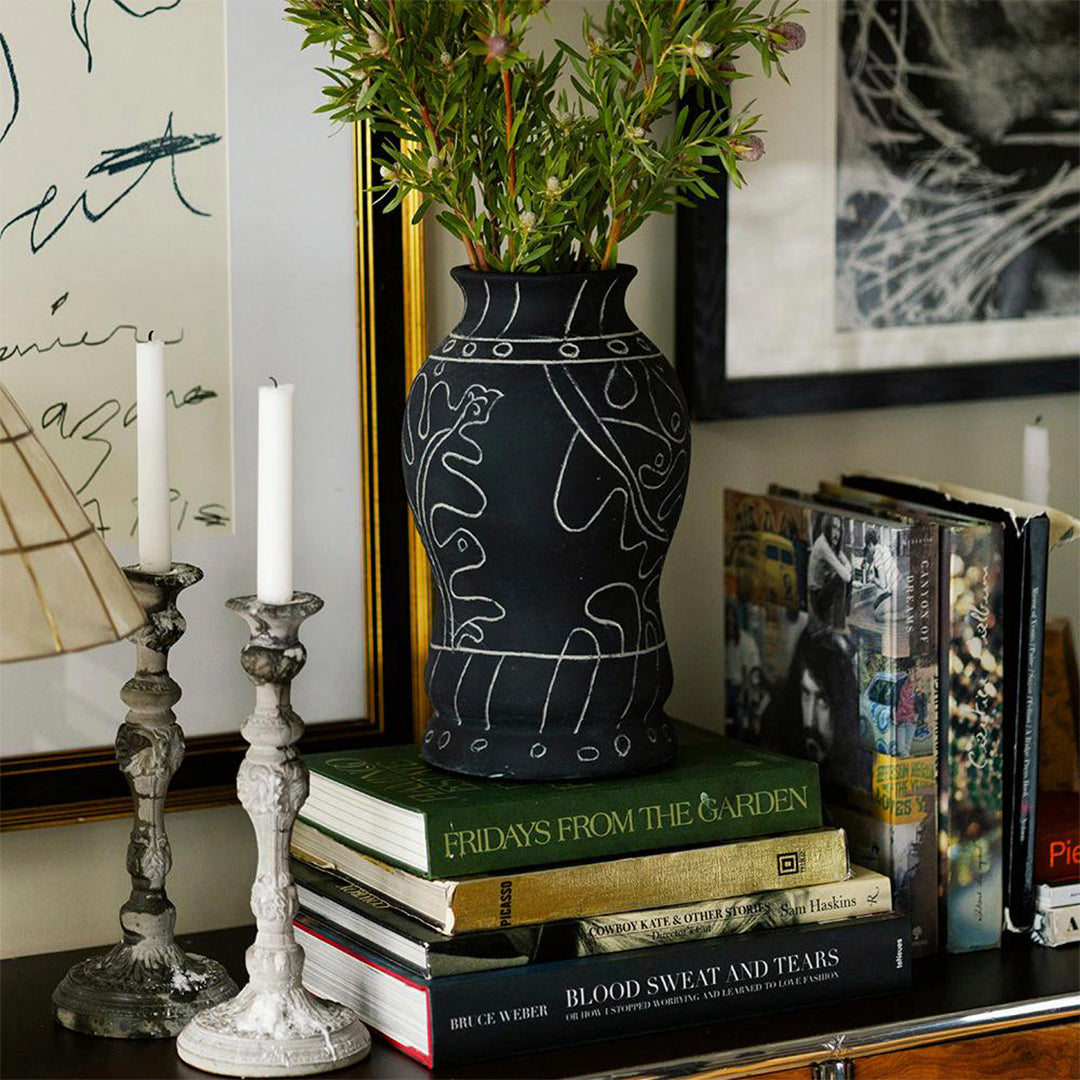 Black vase with white line drawings, surrounded by green foliage, on a stack of colorful books and silver candle holders.