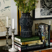Black vase with white line drawings, surrounded by green foliage, on a stack of colorful books and silver candle holders.