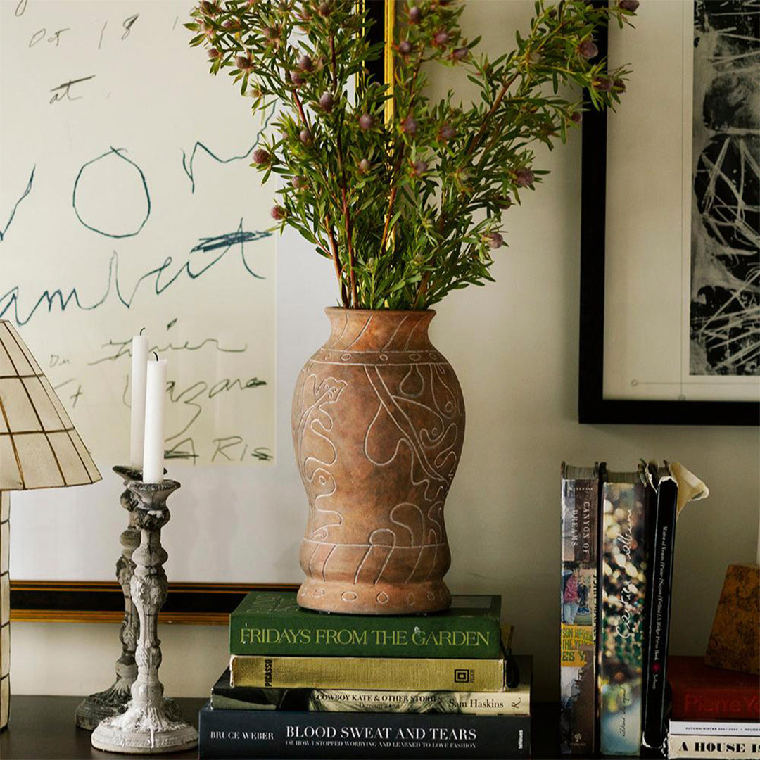 Terracotta vase with intricate white patterns, surrounded by green foliage and layered books in earthy tones.