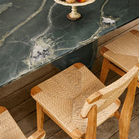 Green marble table with gold veining and woven tan chairs against a wooden floor backdrop.