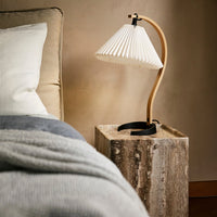 White pleated lamp shade with natural wood base and black accents, complemented by soft gray and cream bedding.