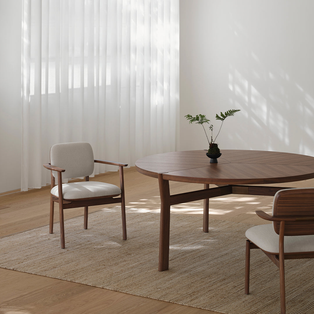 "Light beige armchair on natural wood floor, round walnut table, white sheer curtains, green plant centerpiece."
