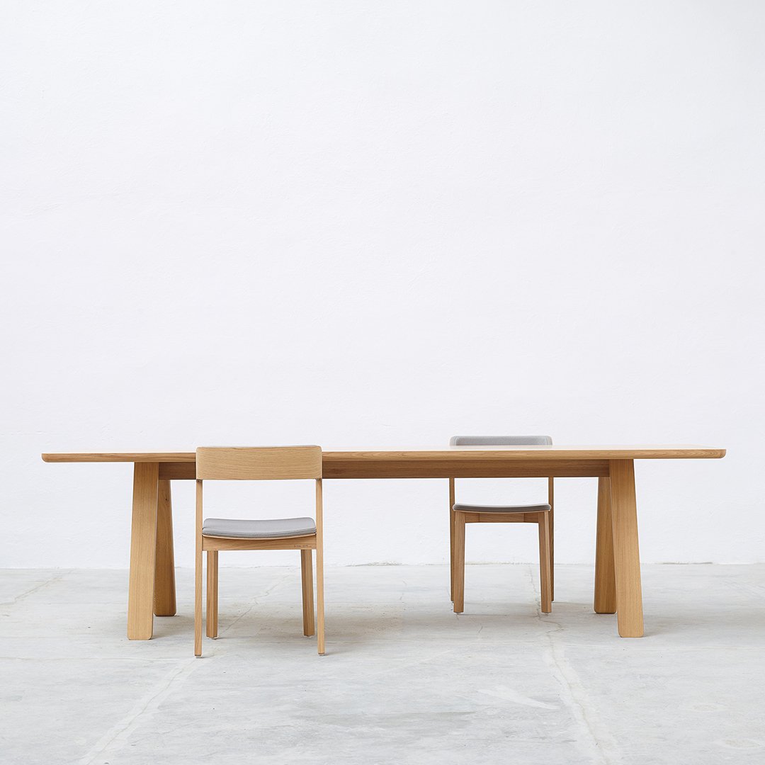 Light oak dining table with minimalist design, complemented by beige upholstered chairs against a neutral background.
