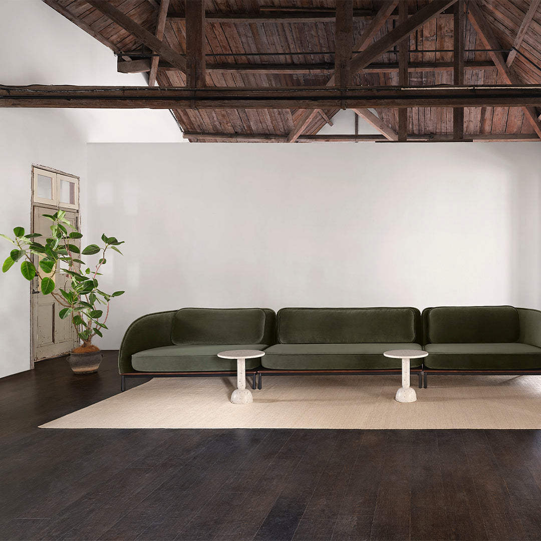 "Green velvet sectional sofa with a minimalist white side table, set against dark wooden flooring and neutral walls."
