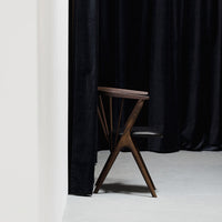 "Contemporary dark wood chair with geometric pattern against a backdrop of black curtains and light gray flooring."