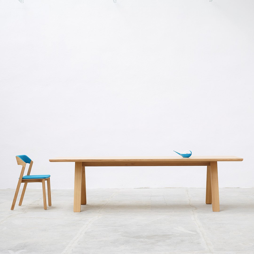 Light wood dining table with a smooth finish, paired with a blue and wood accented chair, against a white background.