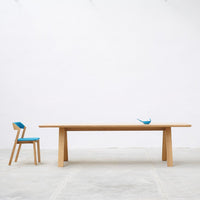 Light wood dining table with a smooth finish, paired with a blue and wood accented chair, against a white background.