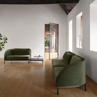 Green velvet sofas with sleek black legs, set against light wood flooring and minimalist white walls.