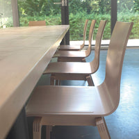 Light wood dining chairs lined along a sleek modern table with a natural finish, complemented by a backdrop of greenery.