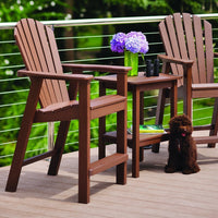 Brown adirondack chairs and table on a wooden deck, set against a green backdrop, with purple flowers as an accent.