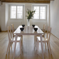 Light wood dining table with four white chairs and black ceramic bowls, set against a bright, minimalist interior.