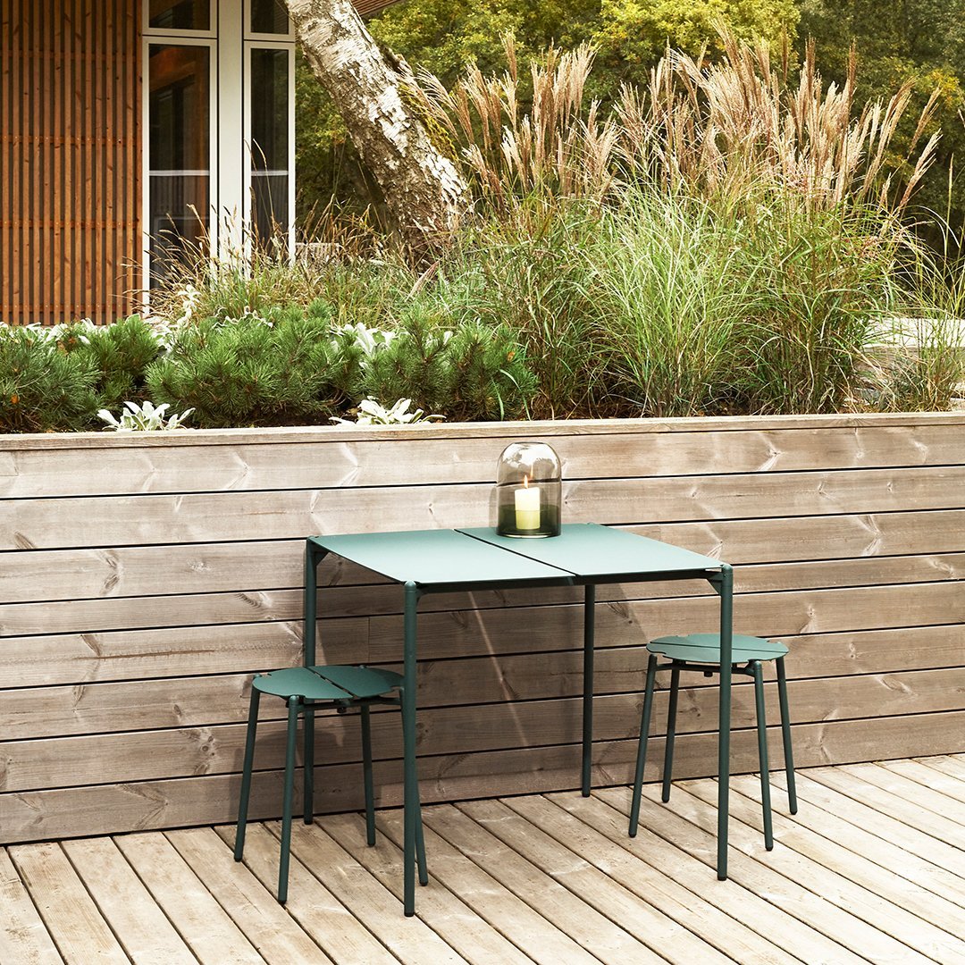 Green metal table and stools with a minimalist design, surrounded by natural greenery and wooden decking.