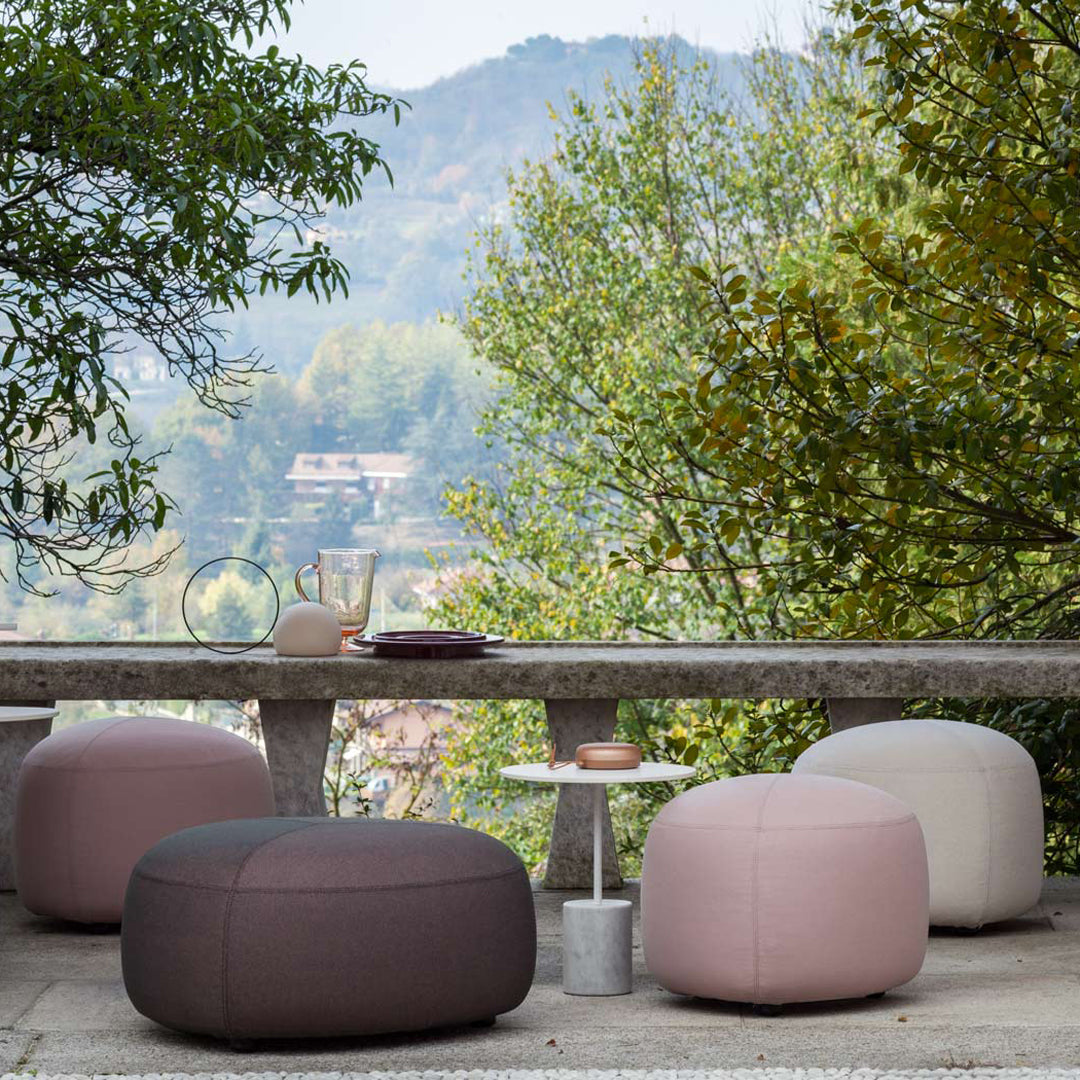 Soft pink, light beige, and dark gray poufs arranged outdoors, complementing the natural greenery and serene landscape.