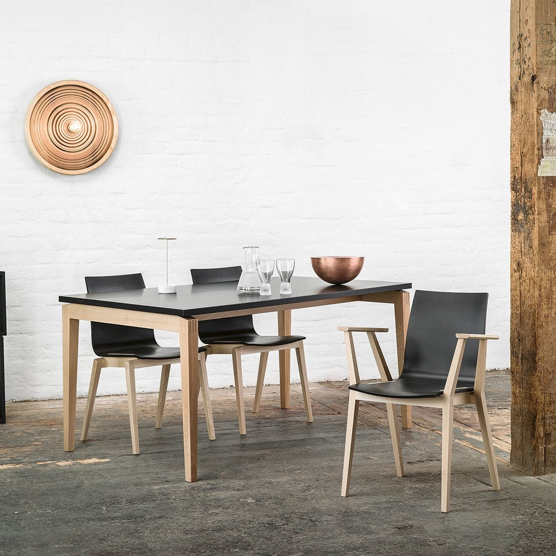 Modern black dining table with natural wood legs and sleek black chairs, set against a white brick wall and rustic floor.