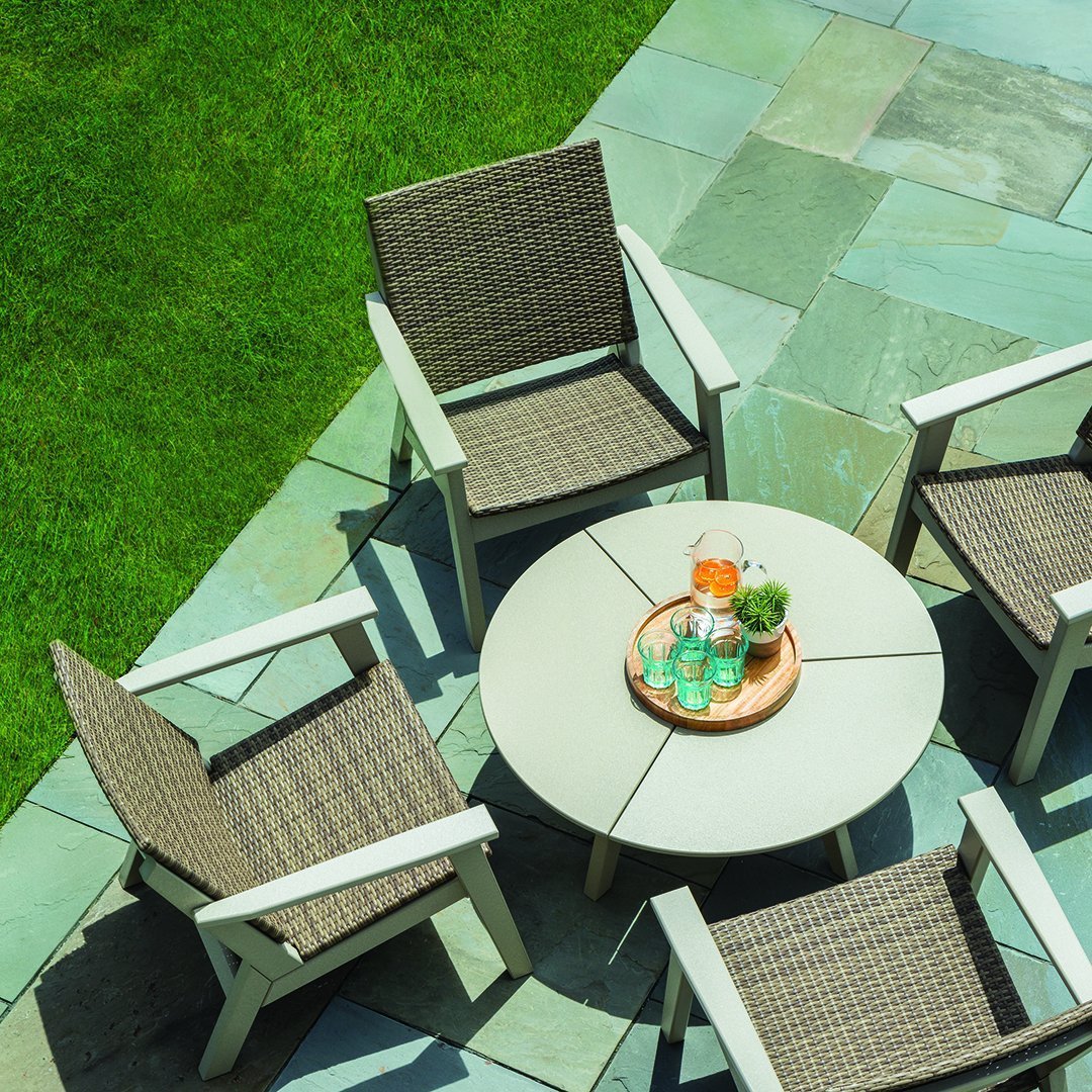 "Neutral wicker chairs with a round white table on a green lawn, surrounded by textured blue and grey stone tiles."