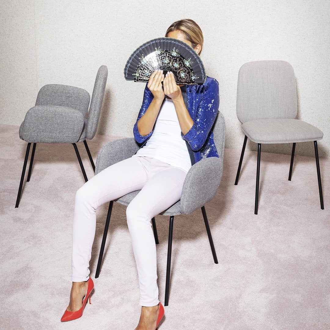 Woman in a blue sequined jacket and white top, holding a fan, seated on a grey chair with pink flooring.
