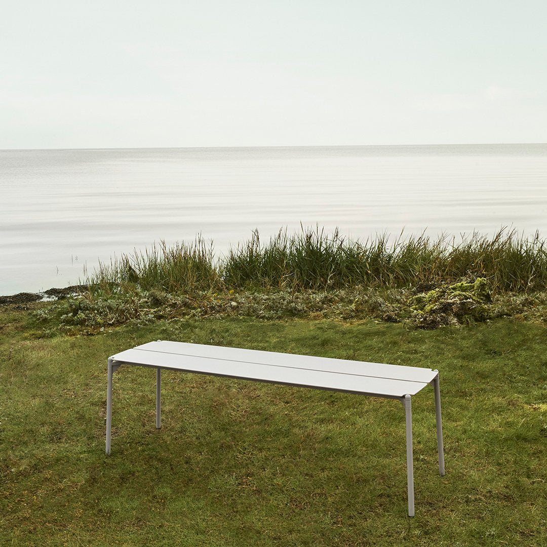 Sleek white outdoor bench with a minimalist design, set against a serene green landscape by the water.