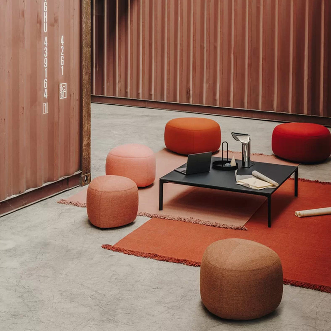 "Colorful poufs in soft pink, orange, and red hues, set on a textured, warm-toned rug with a sleek black coffee table."