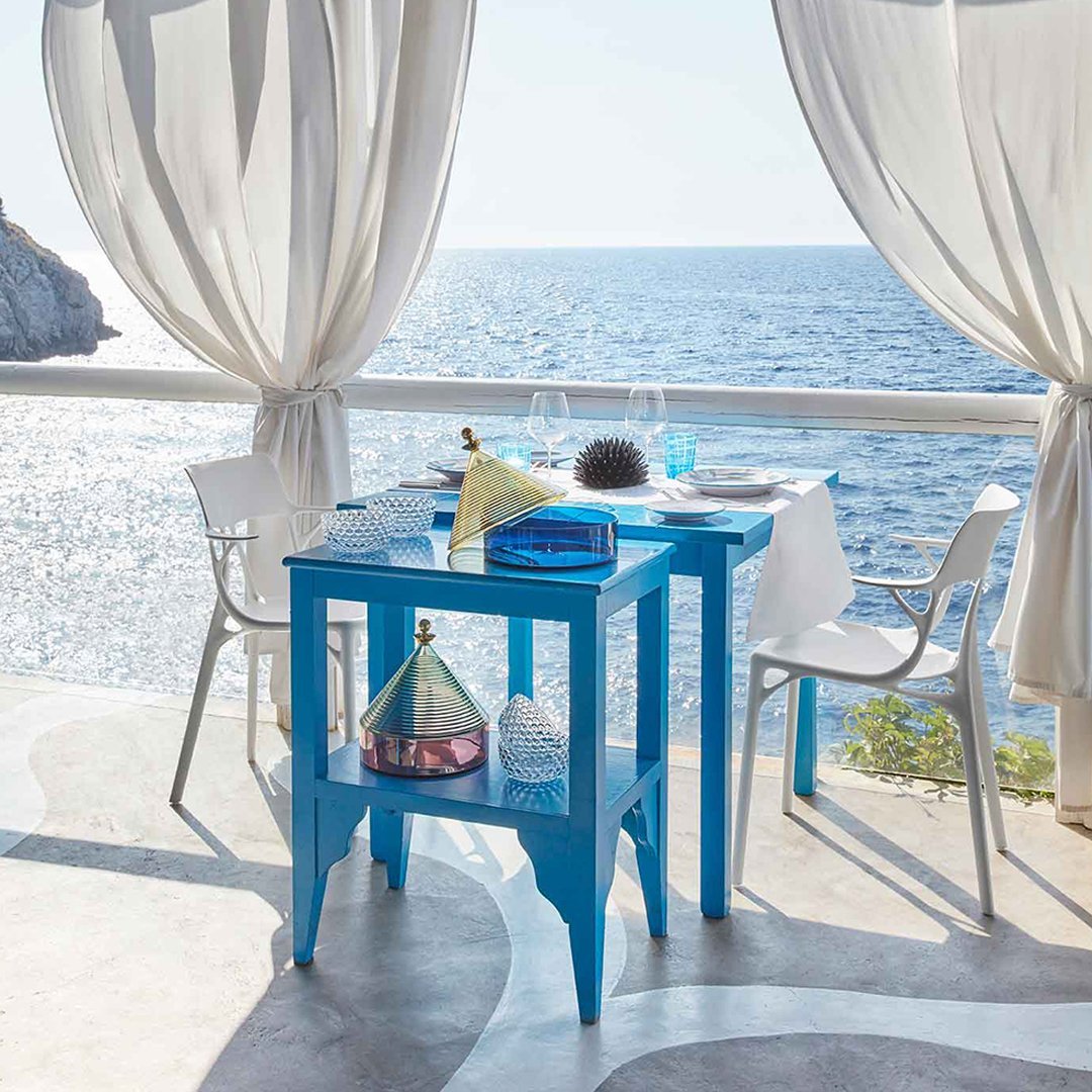 Vibrant blue table with white chairs, set against a coastal backdrop and light, flowing curtains.