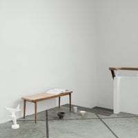 Wooden bench with light brown finish, set against a minimalist gray floor and white walls. Modern decor accents add contrast.