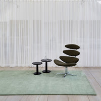 Green modern rug, sleek black tables, and olive green chair against a soft, textured white backdrop.