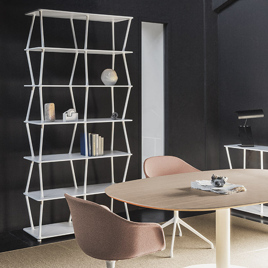 White geometric shelving unit with a black wall backdrop and a round light wood table, complemented by soft pink chairs.
