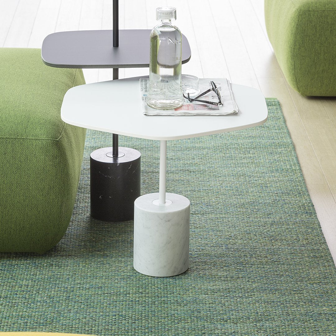 Modern side table with a white top, black and gray base, on a green textured rug and surrounded by a green sofa.