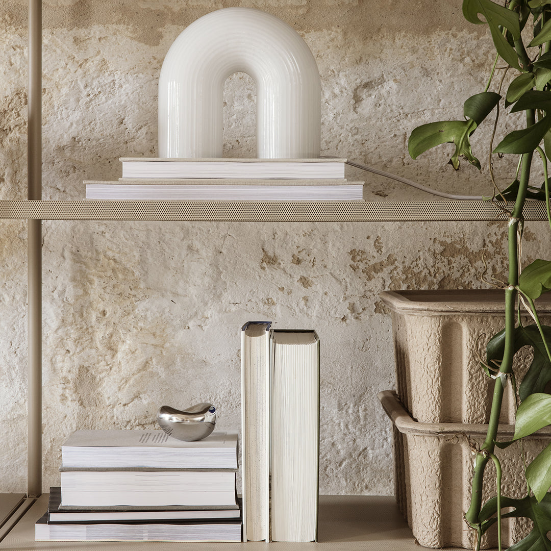 White arch lamp on neutral shelves with stacked books and textured beige planters. Green plant accents.