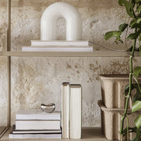 White arch lamp on neutral shelves with stacked books and textured beige planters. Green plant accents.