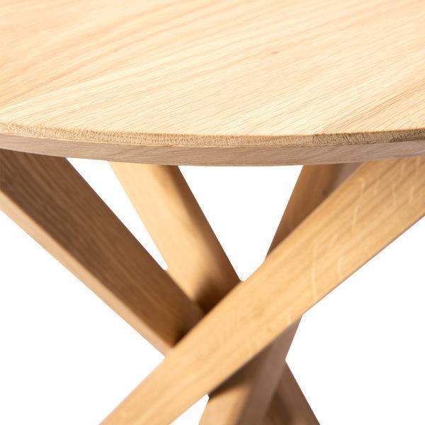 Light oak round tabletop with natural grain, complemented by minimalistic crossed wooden legs in a warm finish.