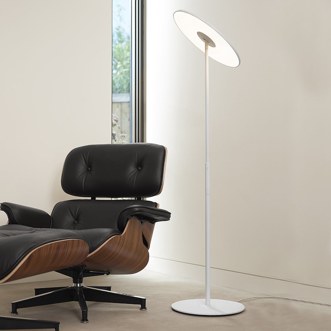 Modern white floor lamp with a sleek circular shade, paired with a black leather chair and wood accents.