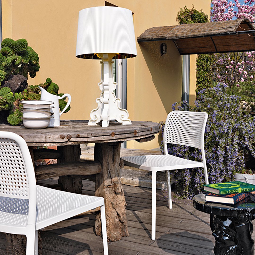 "White woven chairs and a sleek white lamp against a rustic wooden table, complemented by greens and purple blooms."