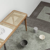 Wooden bench with natural cane pattern on a muted green and gray textured rug, featuring minimalist bowls in black and white.