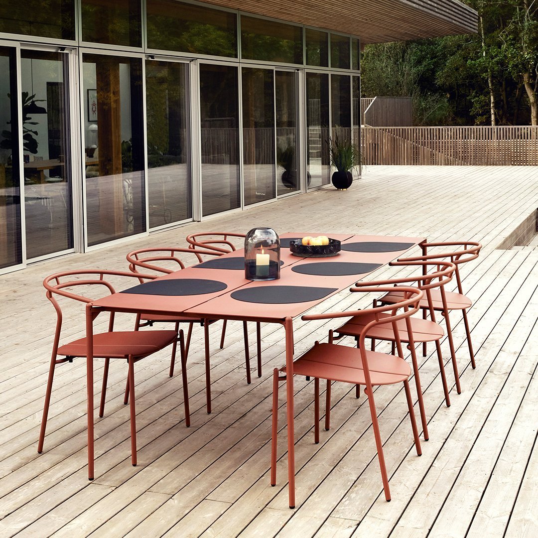 Rustic orange outdoor dining table with round black placemats, surrounded by matching chairs on a wooden deck.