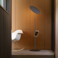 Modern black floor lamp with circular shade, against a warm wood-paneled wall and soft beige floor.