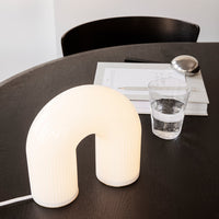 White arch-shaped lamp with ribbed texture on a dark wooden table, featuring neutral colors and minimalist design.