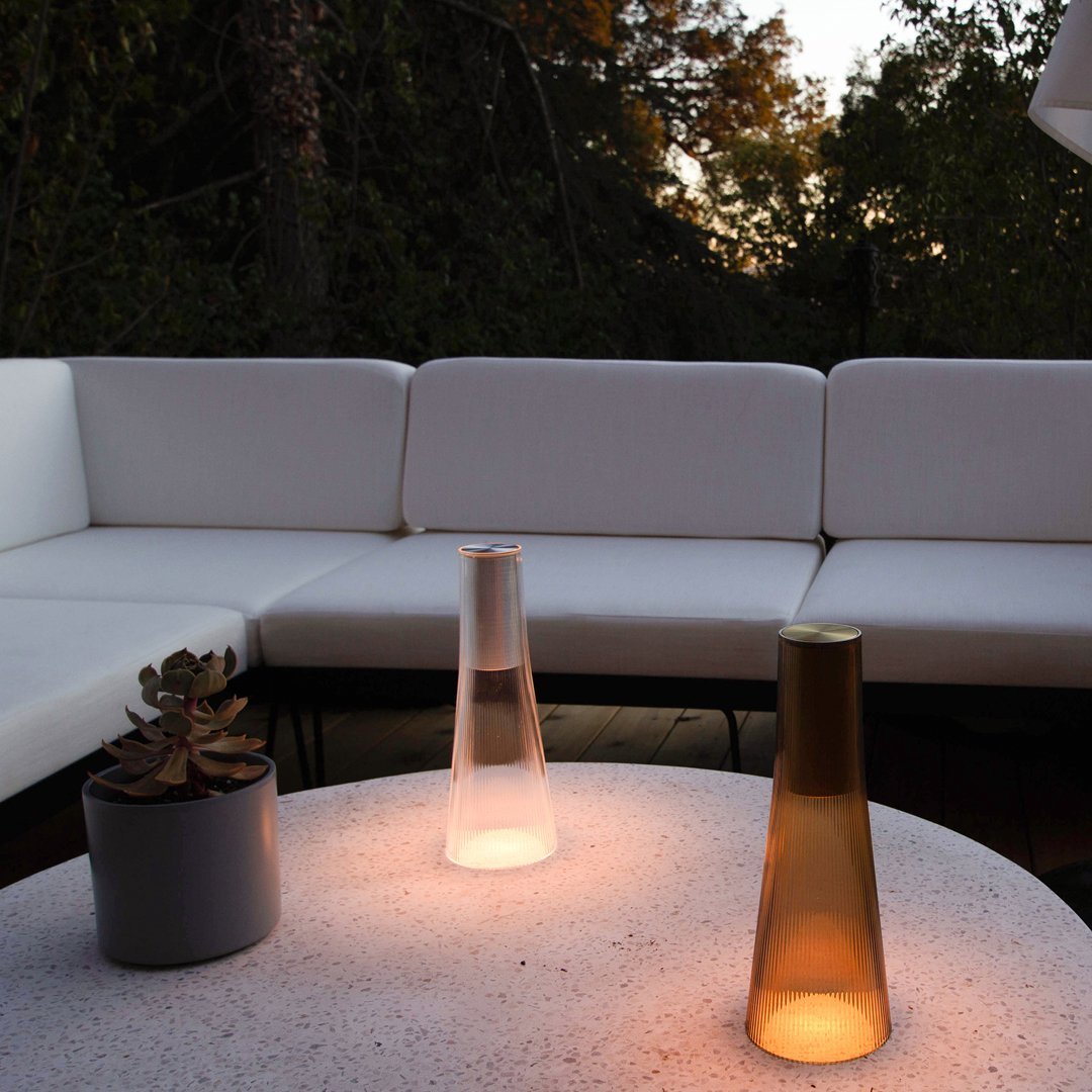 Two sleek, illuminated table lamps in clear and amber glass on a light gray table, with a white sectional couch background.