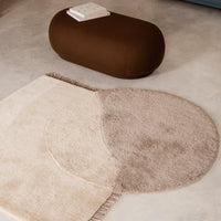 Beige and taupe circular rug with soft textures and fringe detailing, paired with a smooth brown ottoman.